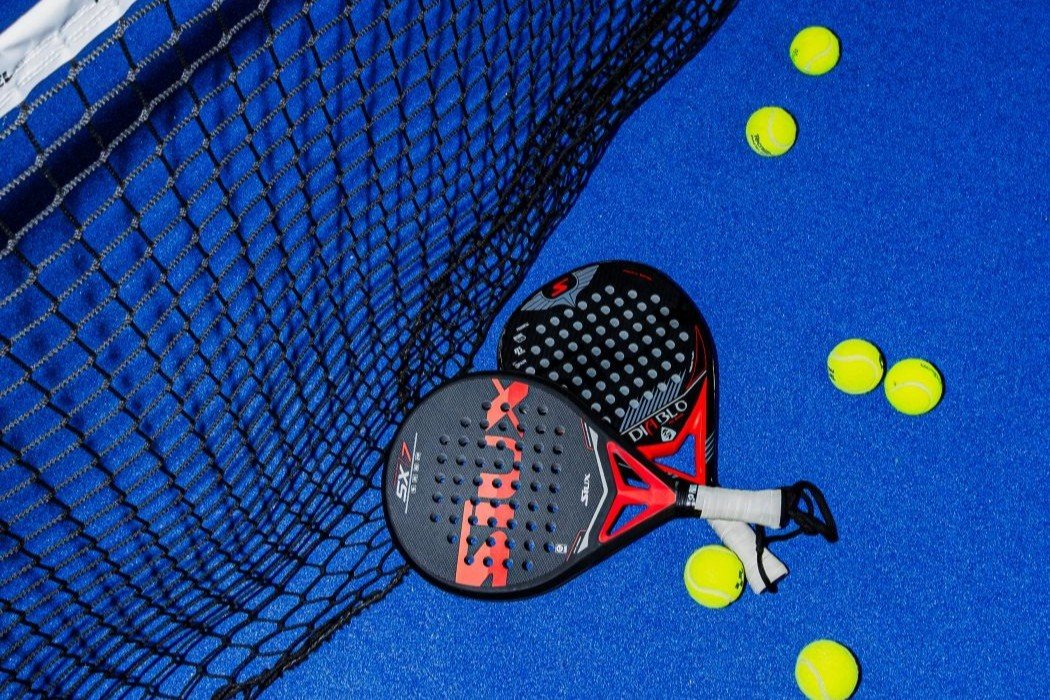 Twee padelrackets tegen het net op de blauwe baan, met gele ballen eromheen